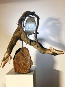 Sculpture made of aspen root of a faceless ballerina in and grand jete withe her hair curing around with a handmade video screen with a projector projecting an AI video made by the artist showing and aged ballerina dancing through an aspen forest who turns in to wood.